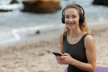 Attractive caucasian woman yogi listening to music with headphones and uses smartphone, relaxing while sitting on mat at the beach