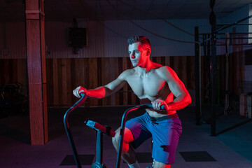 Muscular man training on air bike in red-blue neon light. Cross training