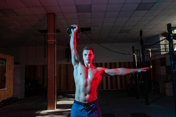Muscular Caucasian man with naked torso training overhead with kettlebell in red blue neon gradient...