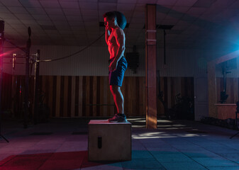 Muscular man training wooden box pacing with a heavy bag in dark gym. Athlete workout in red blue neon light. Healthy lifestyle cross training
