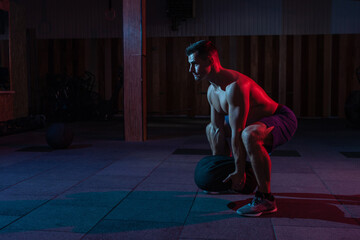 A muscular powerful man with a naked torso exercises with a heavy bag in a red blue neon light....