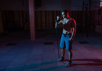 Fototapeta premium Muscular powerful man with naked torso trains with expander (fitness rubber bands) in red blue neon light. Cross workout in modern gym