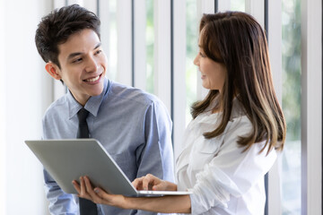 Obraz premium Beautiful black long hair Asian businesswoman standing watching the screen of laptop notebook computer and talking with businessman with intimate manner in office