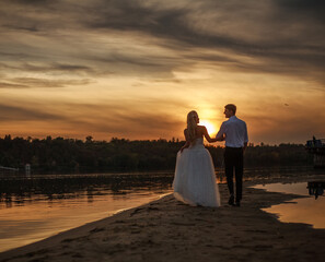 Fototapeta premium Young married couple walk together on a sandy beach, on background of dramatic sunset sky over green forest