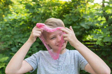 Closeup view photo portrait of cute funny cheerful face of white kid playing cheerfully outdoor in summer backyard and looking through circle made of transparent pink slime toy