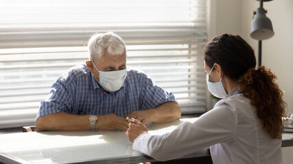 Close up female doctor consulting smiling older patient wearing medical face mask about health insurance agreement at meeting in hospital, therapist and elderly patient discussing contract terms