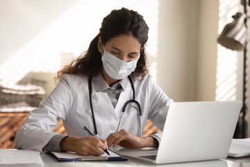 Close up serious professional female doctor wearing protective face mask taking notes, writing in...