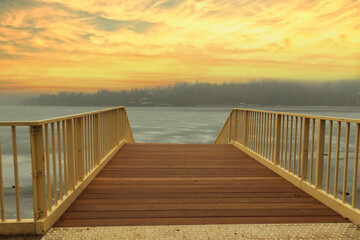 Obraz premium A metal pier with a wooden floor leads to the water. Pier for the steamer Sokolak in the city of Brno in the Czech Republic. There is ice on the water. Sky at sunset.