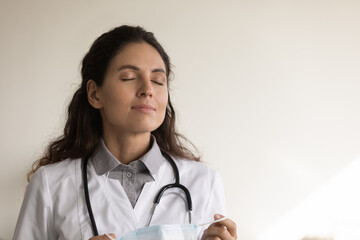 Head shot close up calm female doctor taking off medical protective mask, breathing fresh air deep,...