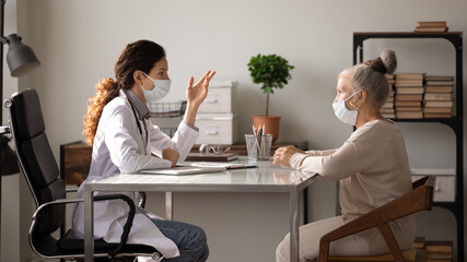 Side view female doctor wearing face mask and uniform consulting mature patient about treatment at meeting in hospital office, therapist and senior woman discussing checkup results, healthcare