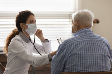 Confident female doctor wearing face mask checking mature man lungs, using stethoscope, listening...