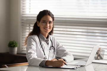 Head shot portrait smiling female doctor wearing glasses and uniform filling medical documents, patient illness history, sitting at desk with laptop in hospital office, student taking notes