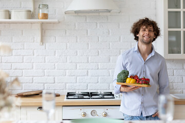 Handsome man with vegetables in casual wear looking at camera and smiling while standing in the kitchen at home.