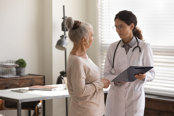 Doctor wearing uniform and glasses consulting mature patient, holding clipboard with medical...