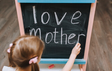 The red-haired schoolgirl painstakingly writes beautifully on a wooden black board, easel, holding white chalk, the words of love for her mother. Photography, copy space, concept.