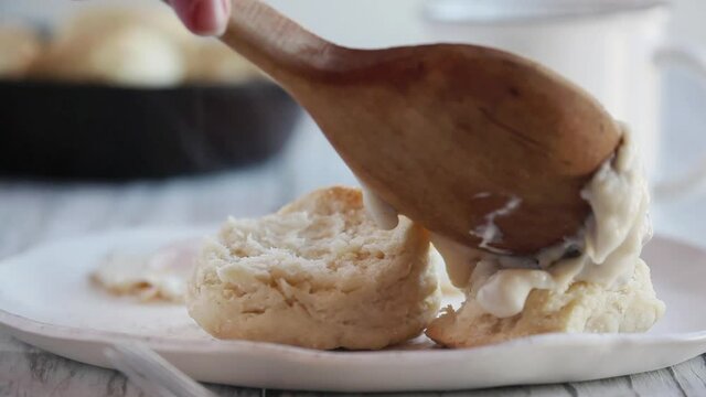 Closeup of woman's hand using a wooden spoon to pour hot steaming gravy over southern buttermilk biscuits.