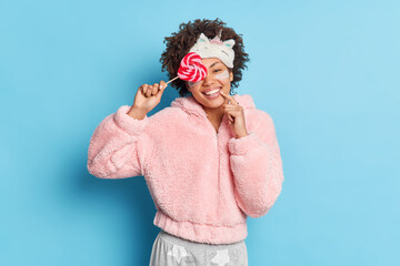 Smiling curly haired young woman covers eye with lollipop smiles broadly dressed in pajama sleepmask isolated over blue background. Beautiful female gets ready for sleep uses skin care products.