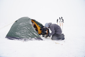 Guys near the tent in a snowy brownstone are preparing food during a winter trip.