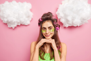 Pretty dark haired young woman with hair curlers makes hairstyle applies collagen patches under eyes to reduce puffiness smiles pleasantly isolated over pink background with fluffy clouds above.
