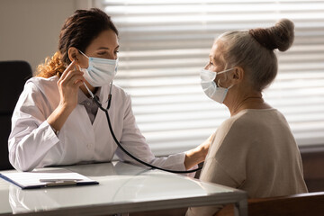 Close up female doctor wearing face mask checking mature patient lungs, holding using stethoscope,...