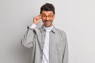 Studio shot of cheerful handsome European guy looks happy through spectacles dressed in grey shirt smiles carefree poses indoor. Happy male employee ready for next task. Positive emotions concept