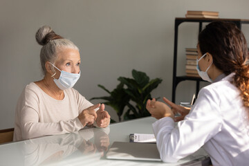 Obraz premium Close up mature woman wearing medical face mask listening to doctor during appointment in hospital office, female therapist practitioner consulting older patient about treatment, elderly healthcare