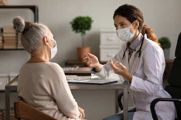 Close up serious doctor wearing medical face mask consulting mature woman patient at appointment in...
