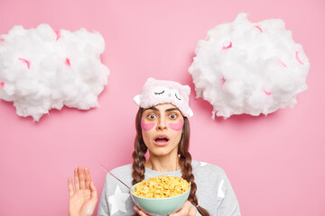 Breakfast time concept. Embarrassed shocked woman with two pigtails keeps mouth opened reacts on something awesome holds bowl of cornflakes dressed in nightwear poses against pink background