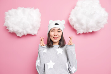 Hopeful smiling gentle Asia woman wears pajama bear hat crosses fingers waits for good news makes wish or prays anticipates something happen isolated over pink background with white clouds above