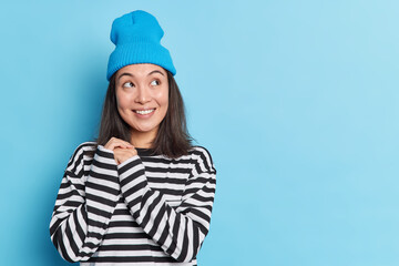 Pretty thoughtful Asian woman keeps hands together looks aside with smile wears hat and striped jumper isolated over blue background with copy space for your promotional content. Pleasant thinking