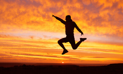 Fototapeta na wymiar Silhouette of a happy man traveler with a raised hand in a jump on a sunset background. Success concept.