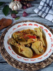 Image of spicy chicken curry or kare or kari or gulai ayam, an indonesian traditional cuisine. Served in enamel bowl over wicker placemat on white wooden kitchen table. Some ingredients appeared. 