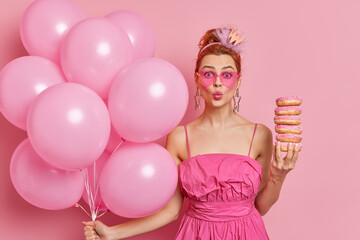 Lovely redhead young woman keeps lips rounded holds pile of appetizing doughnuts celebrates getting new job position poses with inflated balloons against pink background. Festive event concept