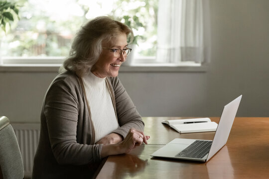 Happy senior retired 60s woman using online app on laptop, looking at screen and smiling. Female pensioner making video call from home, reading good news message, watching funny movie on computer