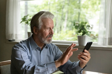 Focused old grey haired 60 - 70s aged man using app on smartphone at home, dialing phone number,...
