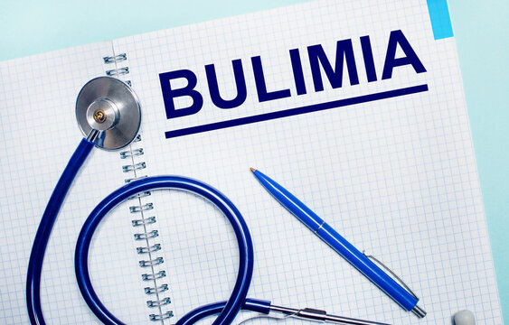 On a light blue background, an open notebook with the word BULIMIA, a blue pen and a stethoscope. View from above. Medical concept