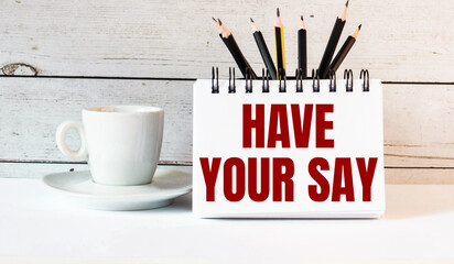 The words HAVE YOUR SAY is written in a white notepad near a white cup of coffee on a light...