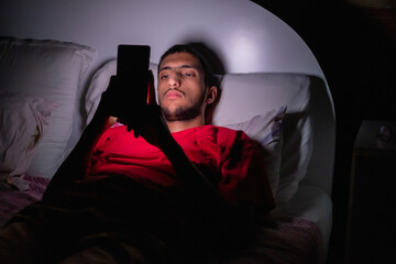 Muslim boy lying on the bed and looking at his phone in a dark room