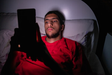Muslim boy lying on the bed and looking at his phone in a dark room