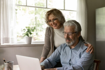 Happy mature OAP couple study internet app on laptop, grandparents use computer at home for online...