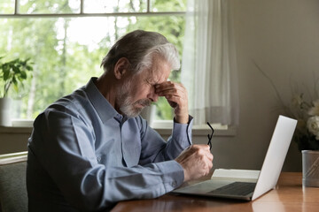 Tired mature 60s man working at laptop from home, taking off glasses, touching head and eyes. Old...