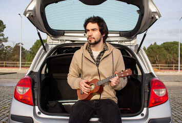 Smiled young man with ukulele in his hands sitting in the trunk. Young man playing ukulele