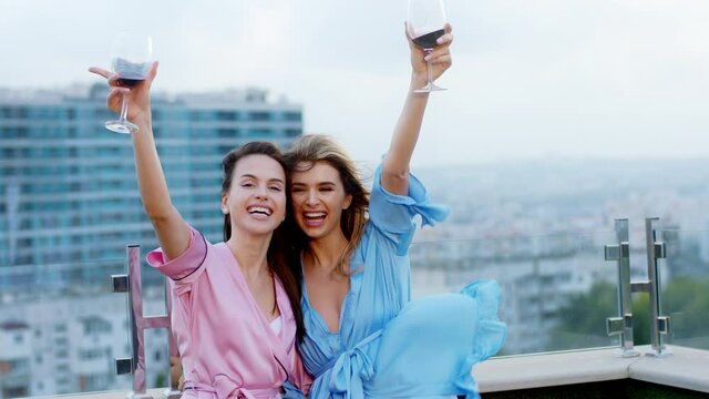 Excited and attractive two best friends ladies in pajamas on the roof top of loft apartment holding glasses of wine and enjoying the evening time