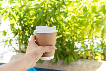 Woman hand holding delivered paper cup of hot coffee on green plants background.