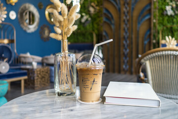 Iced coffee with straw on the white marble table with book and glass vase of dried decoration plant.