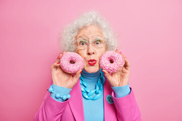 Portrait of good looking grey haired woman keeps doughnuts near face lips rounded enjoys life has sweet tooth wears spectacles fashionable clothes isolated on pink background celebrates anniversary