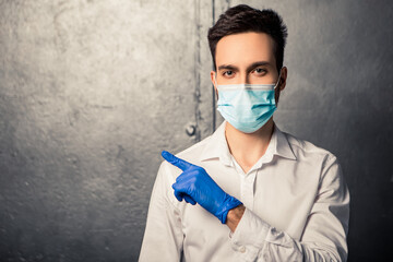 Nurse man pointing in one direction away side copy free space for text message. Doctor dressed in white shirt with blue disposable face mask and gloves standing near the wall.