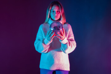Fototapeta na wymiar Young woman holding a disco ball. Girl in a casual sweater on a dark background. She wearing a disco ball. Portrait of a girl student holding a disco ball in hands. Concept - she thinking about party