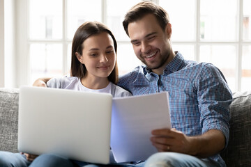 Happy millennial couple reading financial documents at laptop, smiling, hugging. Young husband and wife receiving mortgage loan approval from bank. Homeowners paying small monthly bill online