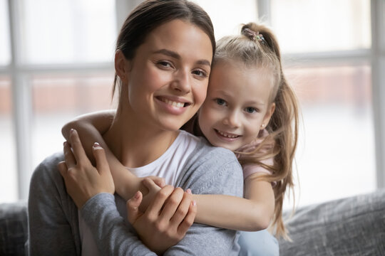Portrait of happy mother and little daughter kid hugging on couch, looking at camera, smiling. Girl and her mon enjoying being at home together, child expressing love and tender to her mum